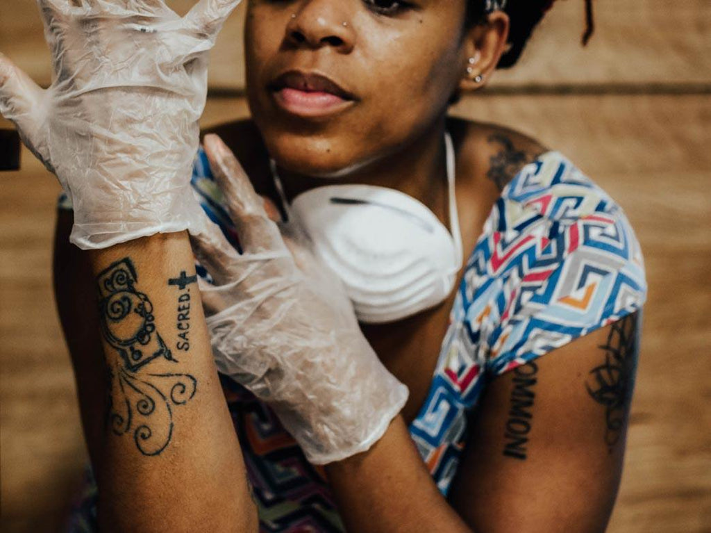 Pandemic highlights racial disparities in U.S. healthcare: close up photo of a Black woman with tattoos on her arm. They are wearing plastic gloves and have a mask around their neck.