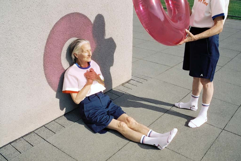 Will a meaningful life help us face death? A photo of an elderly couple. A woman sits against the wall, her face covered in a red halo created by her husband holding a round red plastic beach toy up to the sun.