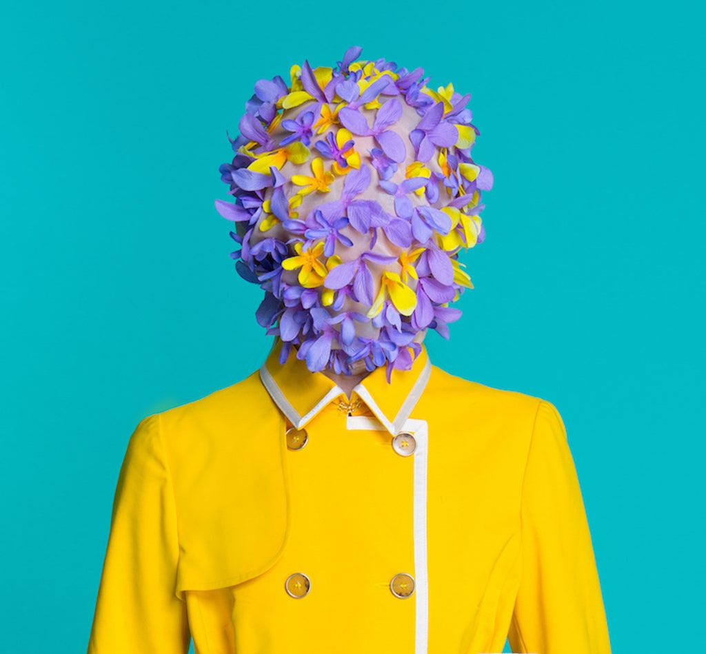 Fashion style model in yellow coat and art accessories on head posing in the studio on blue background purple and yellow paper symbolising living with multiple chronic illlnesses, including brain lesions.