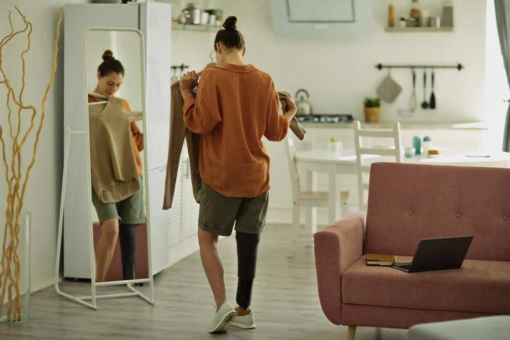 What is adaptive clothing? This is a photo of a woman amputee looking in a mirror while trying on clothes in cozy home interior, copy space.