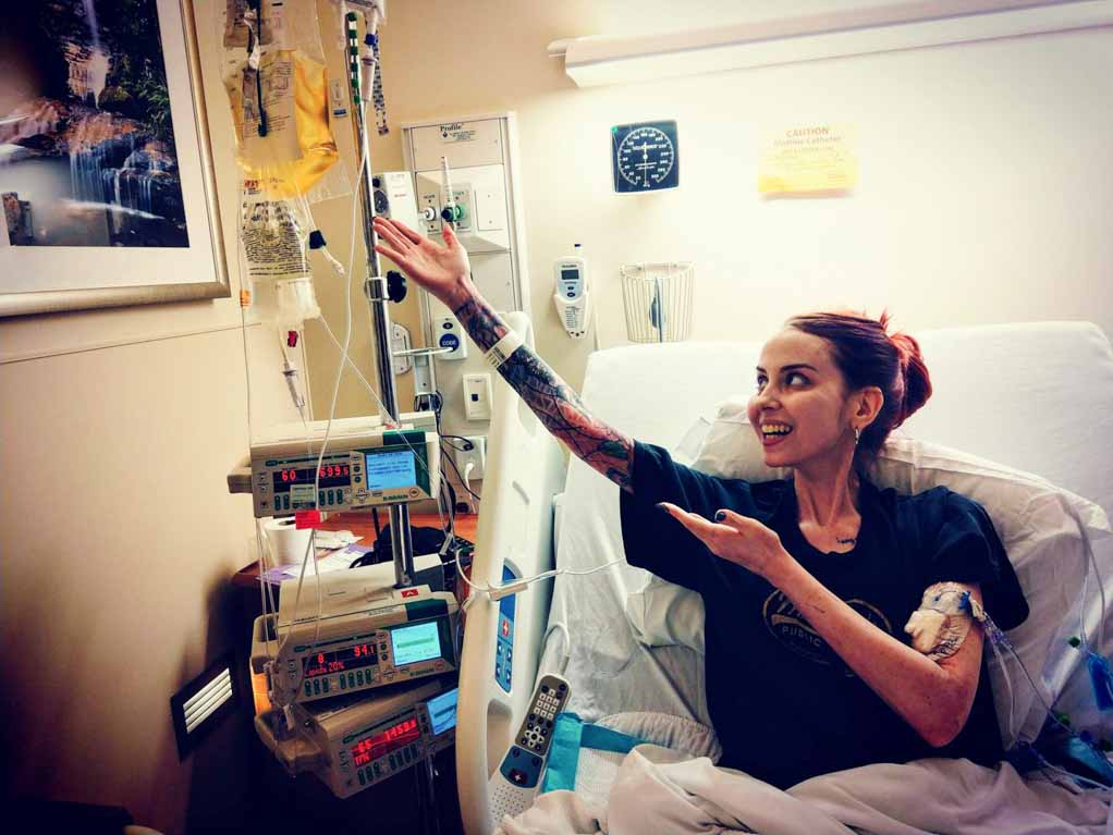 Social media and chronic illness. Photo of Sarah Birdstrong in hospital for Crohn’s Disease sitting up in bed pointing at her IC drip.