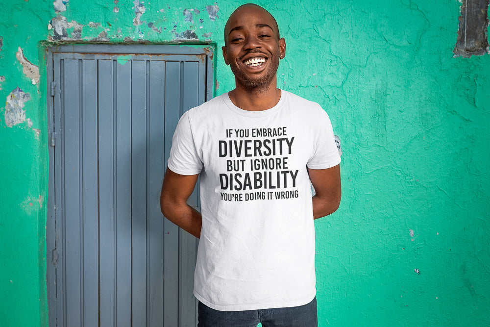 Not all disabilities are visible: a man is standing in front of a teal wall. They are wearing a t-shirt that reads: "If you embrace diversity but ignore disability you're doing it wrong."