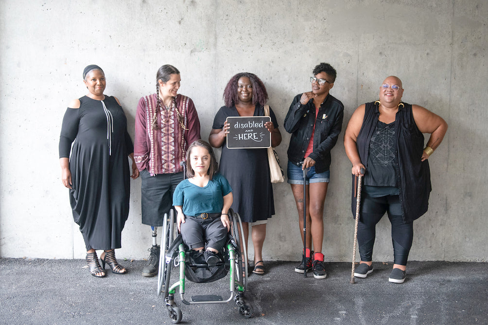 Disability rights movement: six disabled people of color smile and pose in front of a concrete wall. 