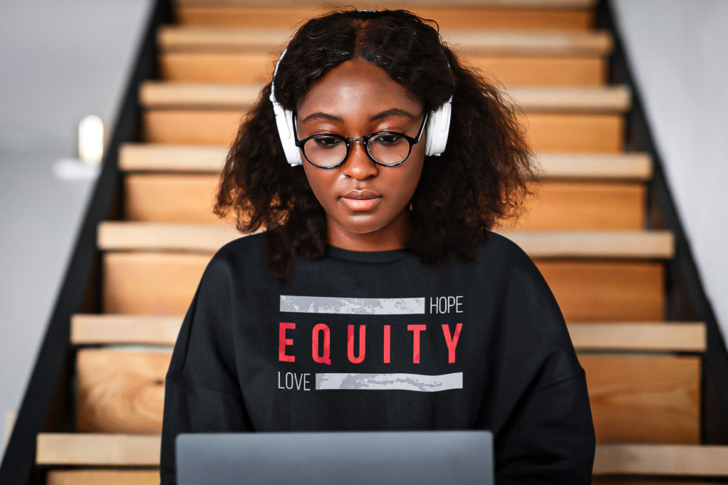 A Black woman is sitting on some wooden stairs wearing headphones at work while working on a laptop.