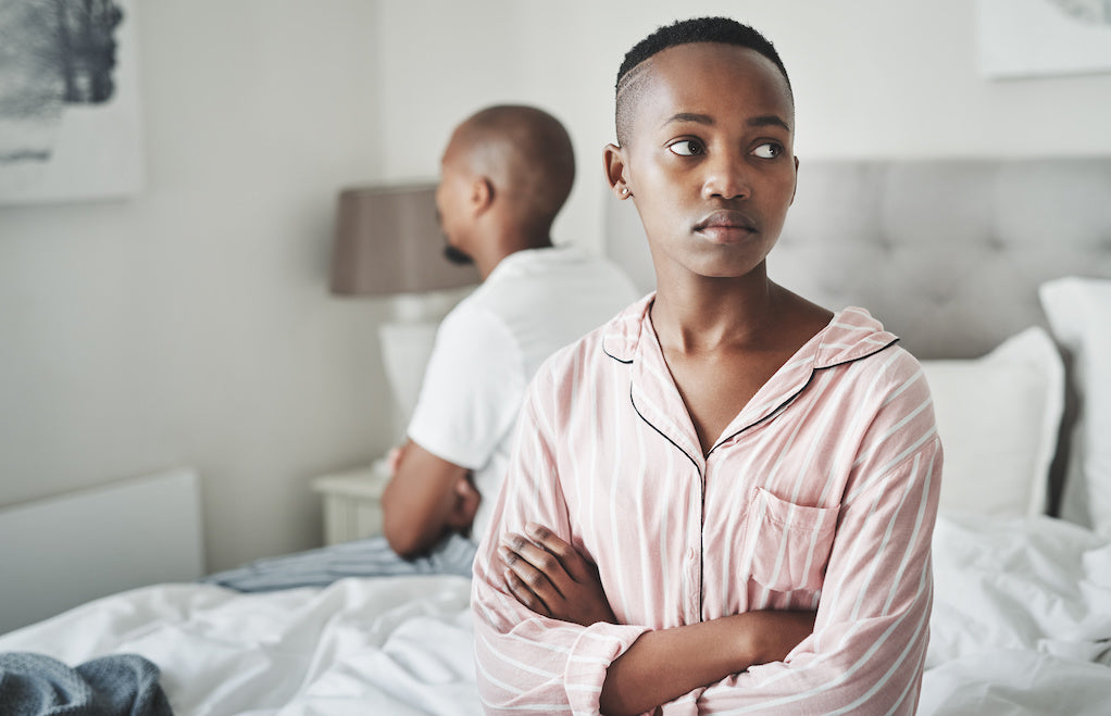 Image for an article explaining what is vaginismus. Young upset black woman is sitting on the edge of the bed, her boyfriend is lying in bed. There is tension because of the impact vaginismus has on their relationship.