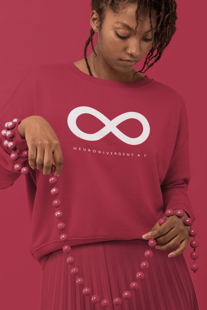 Neurodivergent clothing collection: this is a photo a tall woman is wearing original neurodivergent merch by URevolution. She is wearing red neurodivergent af sweater. She is looking at a set of long red beads in her hand.