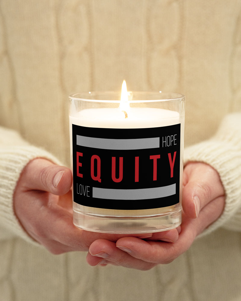 An Equity candle in a glass jar. On the outside of the jar is a black label. The word 'Equity' in upper case red letters is in the middle of the candle. Above and below the word are two thick rectangle blocks colored grey. The word HOPE is printed on the top right-hand side, and the word LOVE is on the bottom left side of the block. The lit candle is being held by a person on front of their chest.