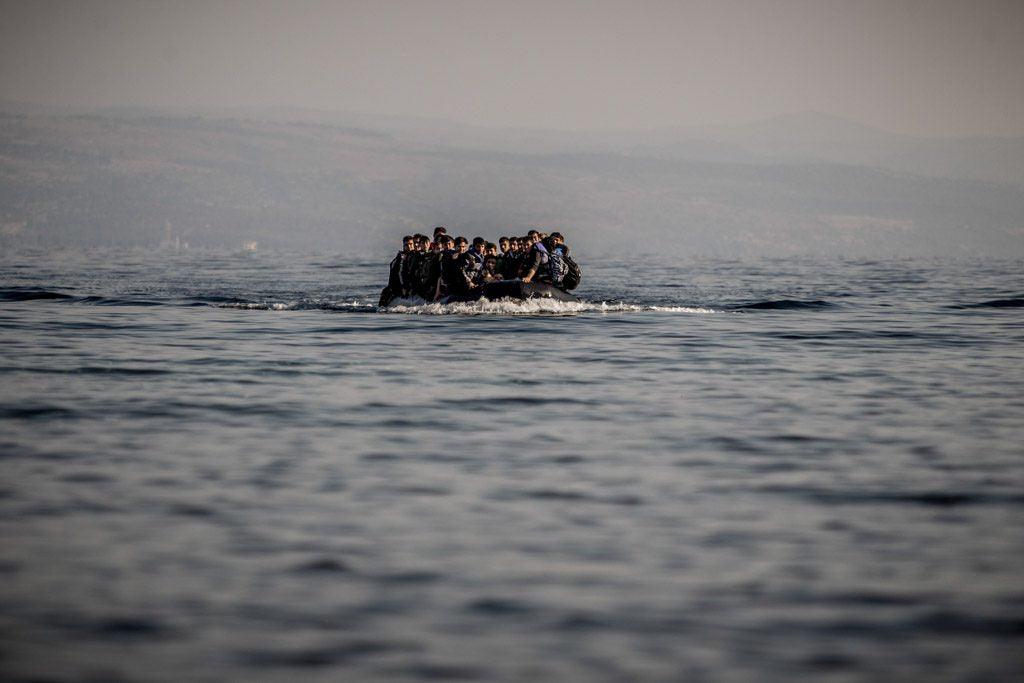 A boatload of migrants in an inflatable boat is at sea, the shadow of shoreline behind them.