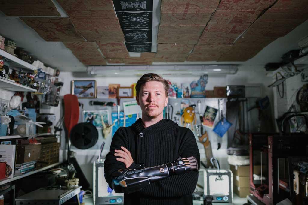 3D printed prosthetic arms: photo of Dan Melville standing in his design workshop with his arms crossed. His 3D printed bionic arm is visible.