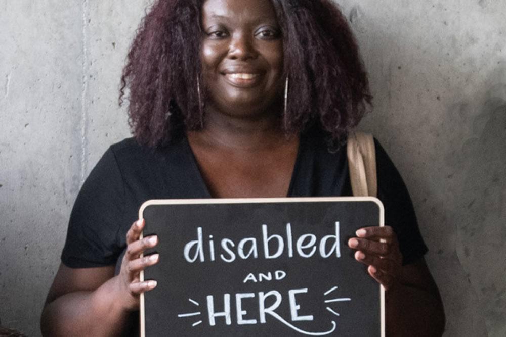 A smiling woman holds a chalkboard sign reading “disabled and HERE.”