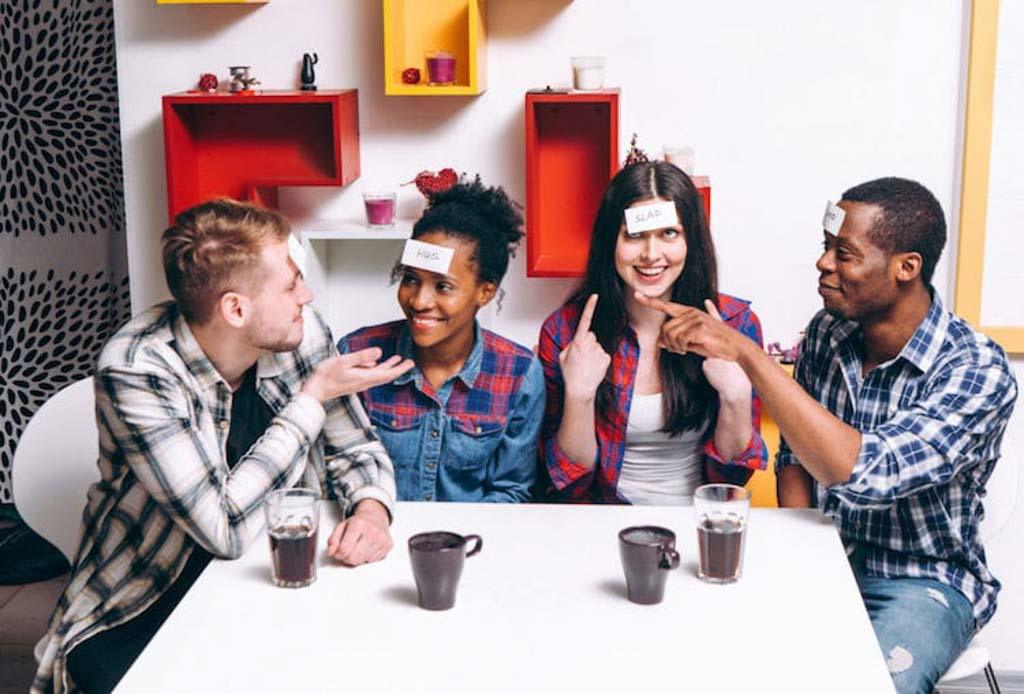 Group of four friends with labels of their medical diagnosis taped to their foreheads all point to one another's labels in an attempt to guess what's written on their heads. 