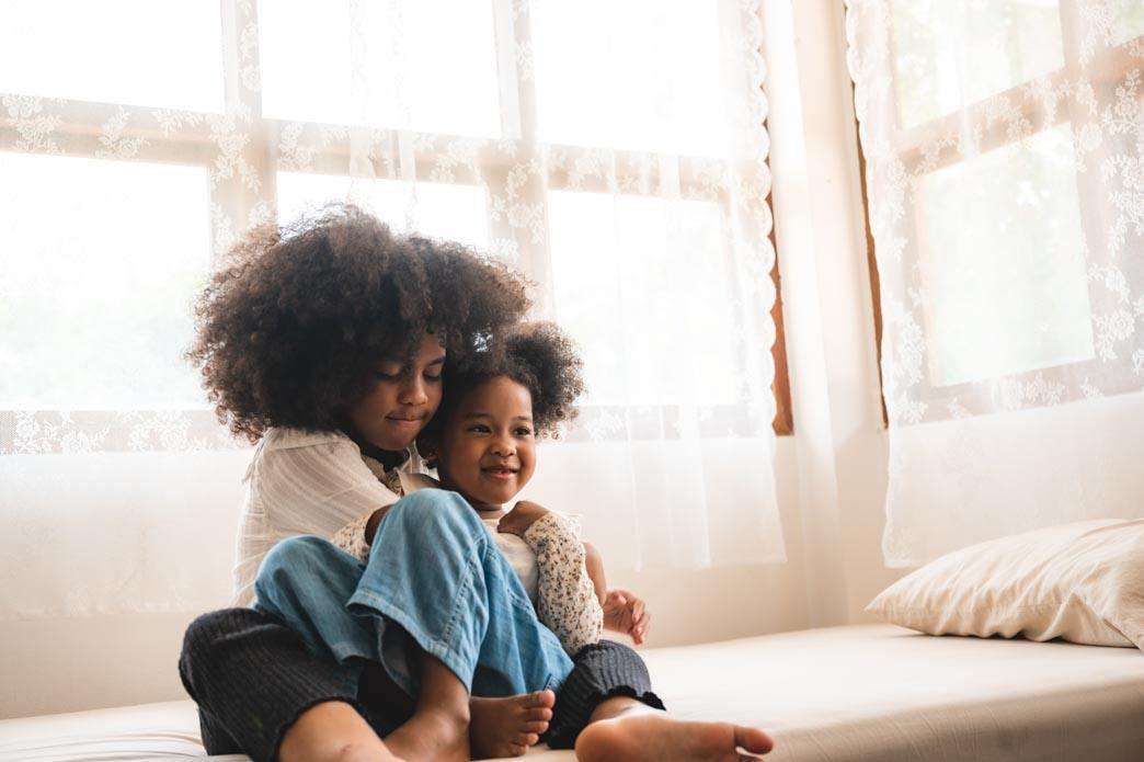 Two children sit together on a sofa, with the older child holding the younger child.