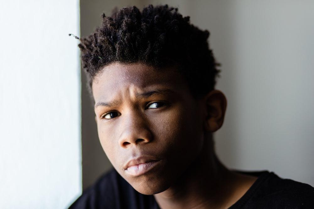 Doctor patient confidentiality for teenagers: why it matters. A teenager looks straight to camera with a strong, concerned expression. He looks vulnerable and his eyes reveal he has a secret to share. He has a spiky afro and is wearing a black t-shirt.