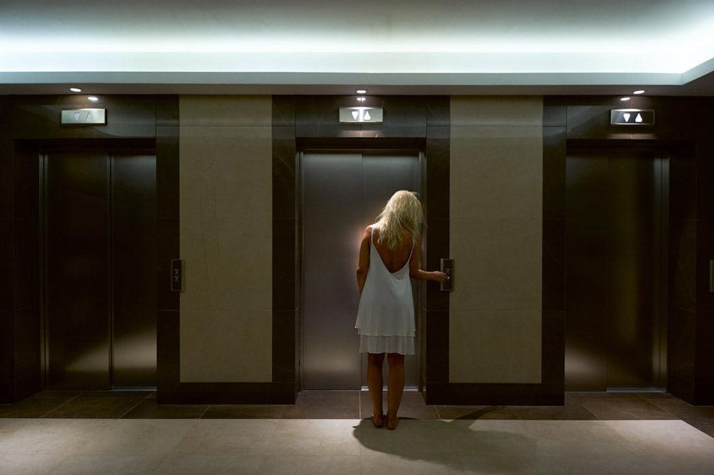 An elevator story: a woman is standing in her nightgown in from of a bank of three elevators. She is in a dimly lit hotel hallway.