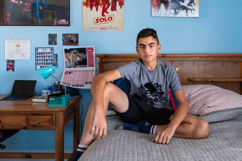Sam Mazaheri sits on his bed in shorts and t-shirt looking at the camera.