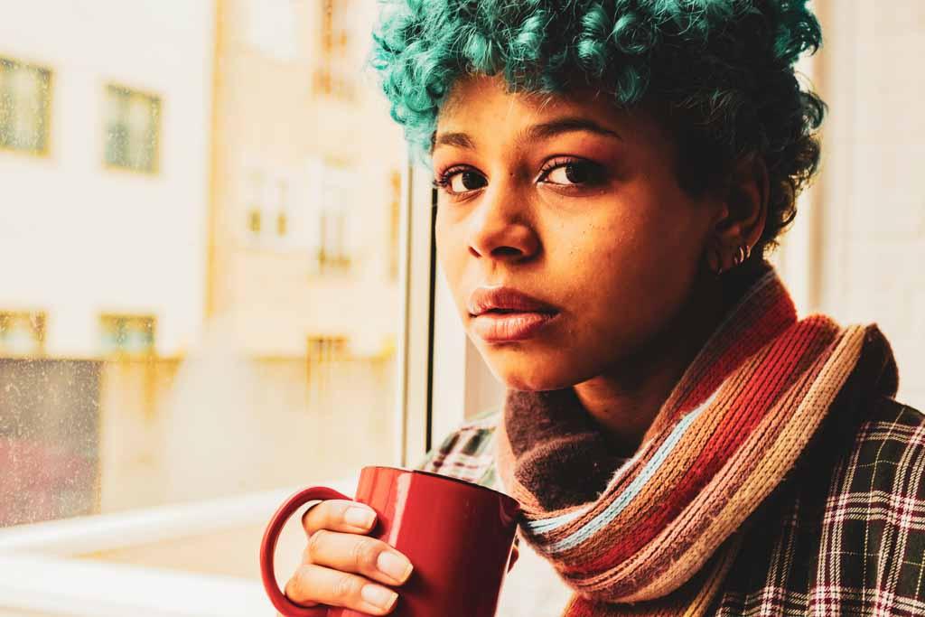 Mental illness in Manhattan: portrait of a person with blue curly hair at the window with a cup, with natural light lighting their face.