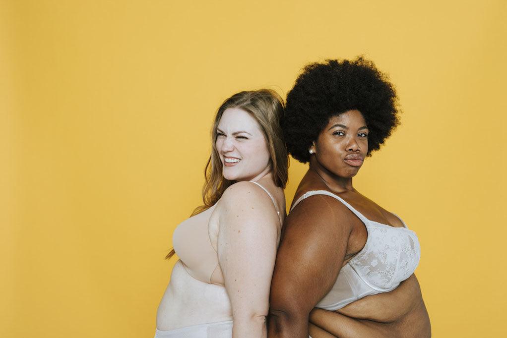 Two women in underwear standing back to back against a yellow background.