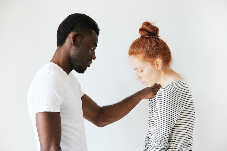Photo of a compassionate man comforting a woman with red hair, his left arm is on her right shoulder, she is slightly huddled forward with sagging shoulders, and her head turned downward. 