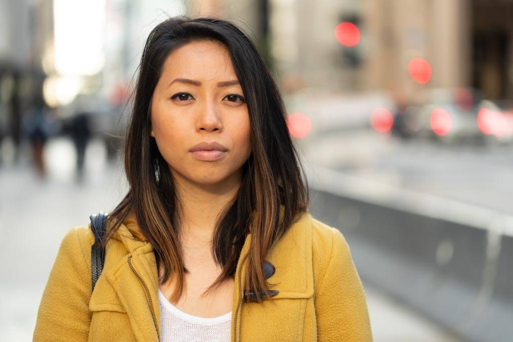 Colostomy bag stories: image of an Asian woman standing on a sidewalk wearing a yellow overcoat. The background is blurred.