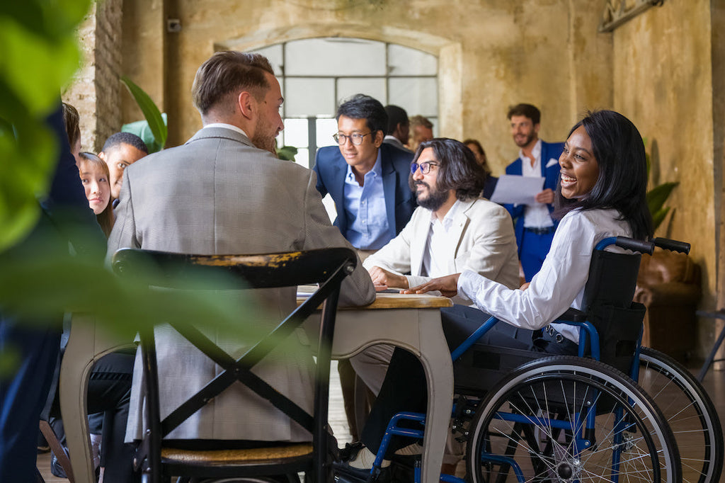 A photo of a multiracial team sitting around a table discussing how to build accessibility into corporate events. The group is multiracial and diverse, including a Black woman wheelchair user having discussions with colleagues