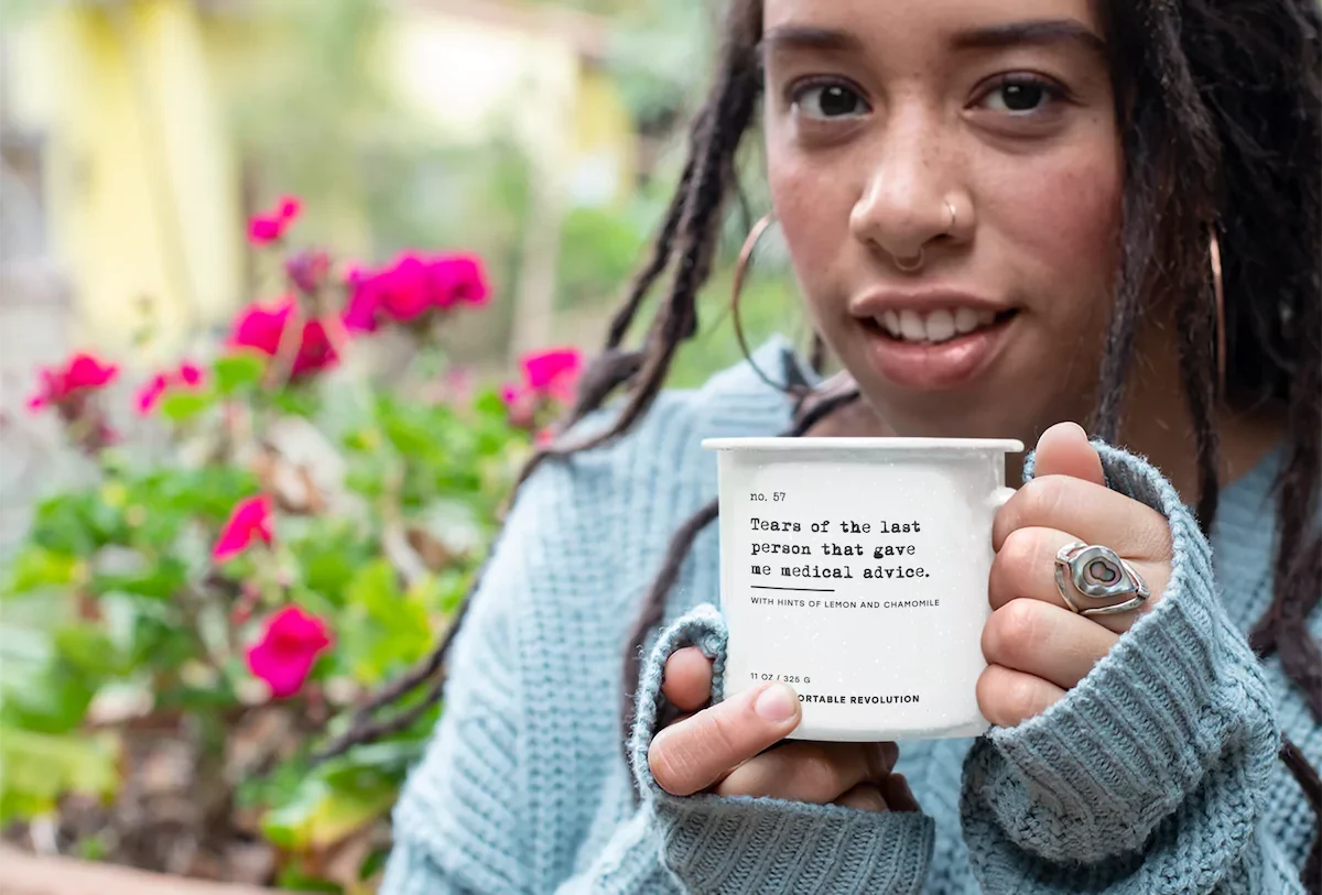 Person with locs holding a white enamel mug printed “Tears of the Last Person That Gave Me Medical Advice” in a garden.