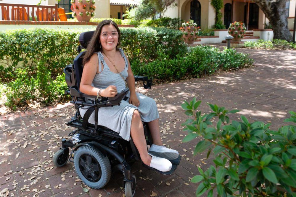 Sylvia Colt-Lacayo, who has a neuromuscular disease, sits in her wheelchair on a. garden path.Image for article on access to college for disabled students.