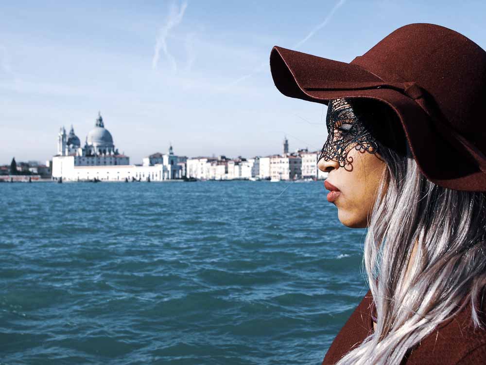 Side profile shot of a young person looking straight ahead having depression on vacation in Venice. They fill up most of the right of the photo. Venice is in the background. 