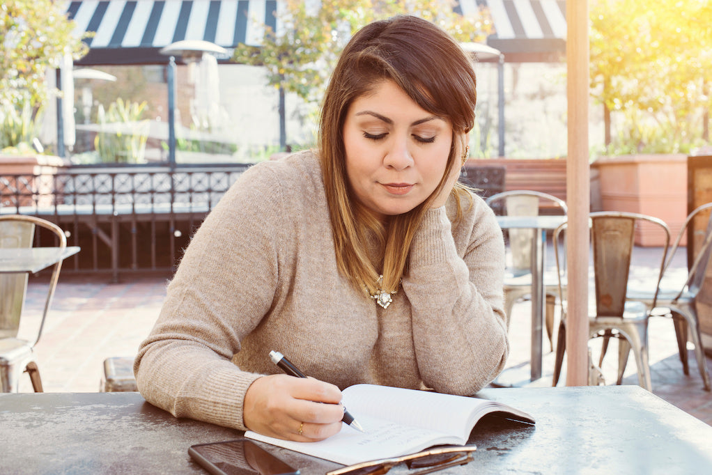 A woman is sitting outside writing in her BPD gratitude journal in a cafe.