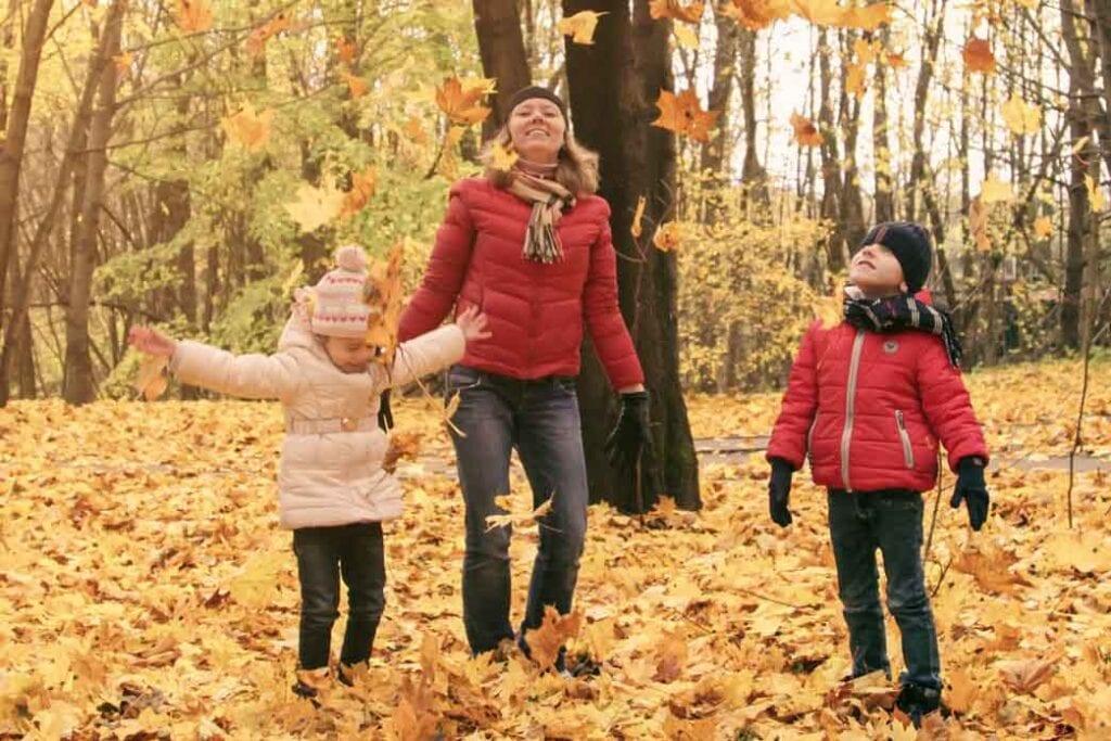 An adult and two children walk among fallen leaves in a park.