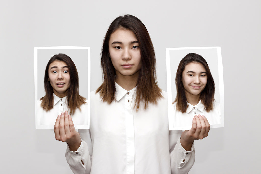Can autistic adults recognize facial emotions? A woman with a neutral expression is standing indoors against a grey wall. They are holding up two photos of themselves: the one on the left is her looking surprise and the one on the right is her smiling.