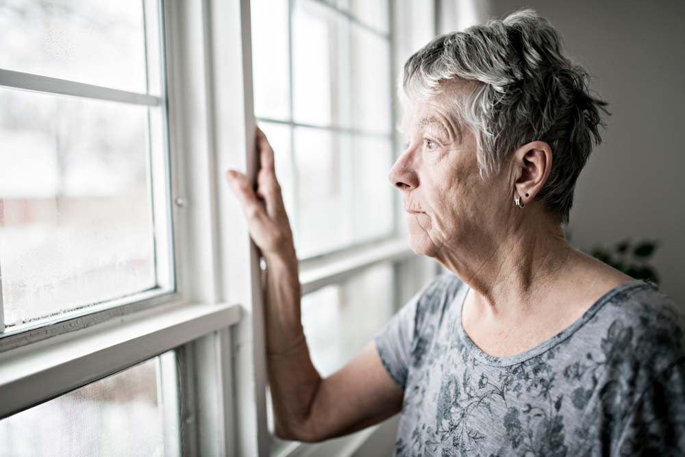 The chronic illness of long covid: an older woman with short grey hair stands next to a window looking outside.