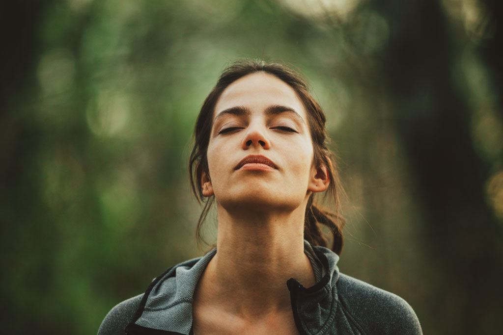 An open letter about depression and illness- photo of a young woman with eyes closed taking a deep breath in a forest.