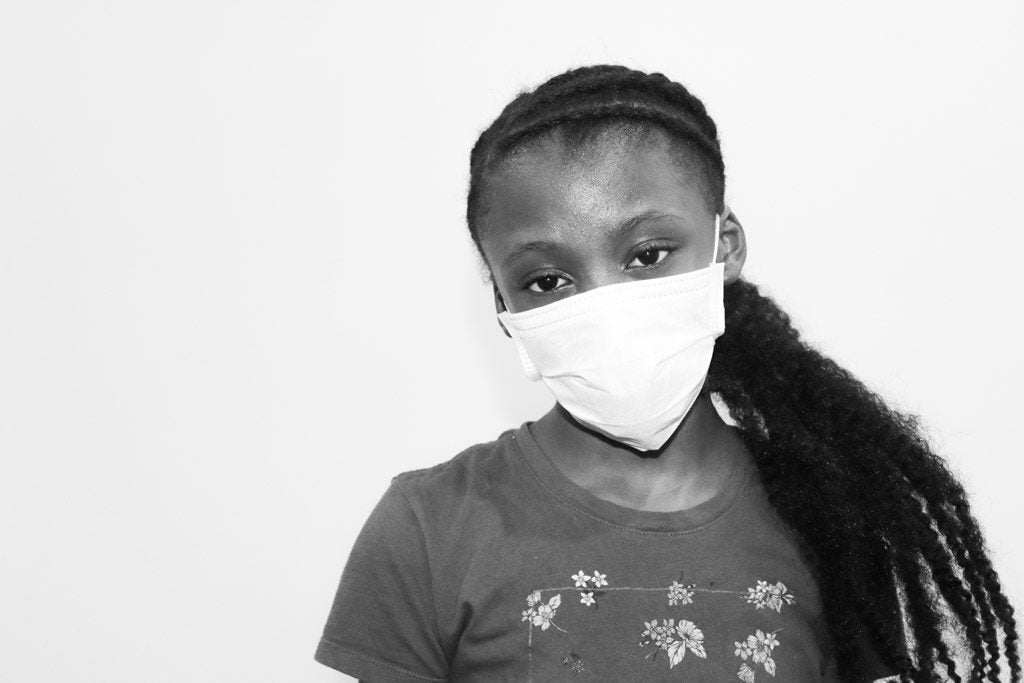 Black and white photo of a young African-American girl wearing blue surgical mask tan background for an article on the coronavirus pandemic and racial status