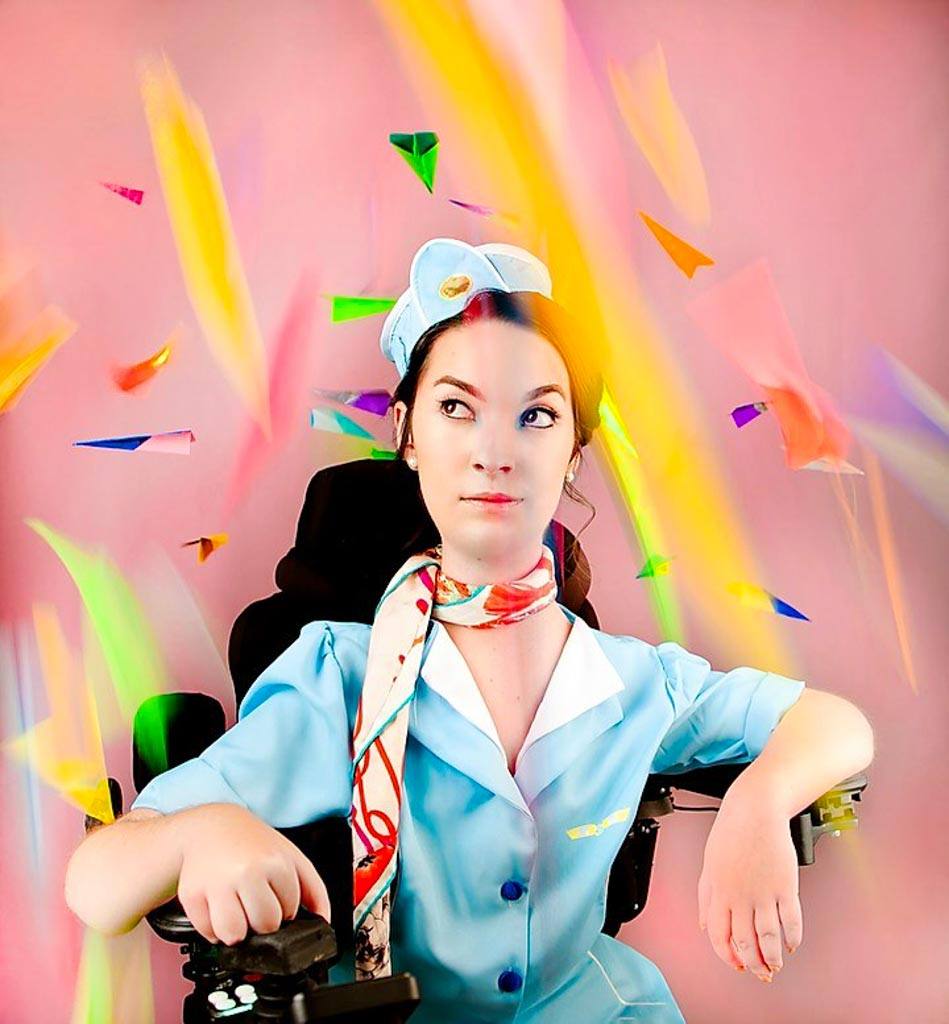 This is a stylized photo Glow Rivera Casanova, sitting in a wheelchair, dressed as a flight attendant. The uniform is light blue, with a retro-style 1950s look. Streaks of colorful paper fall across the photo, giving the appearance of movement.