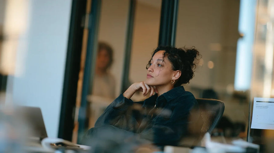 Professional seated at a desk in a modern office, looking toward a meeting room.