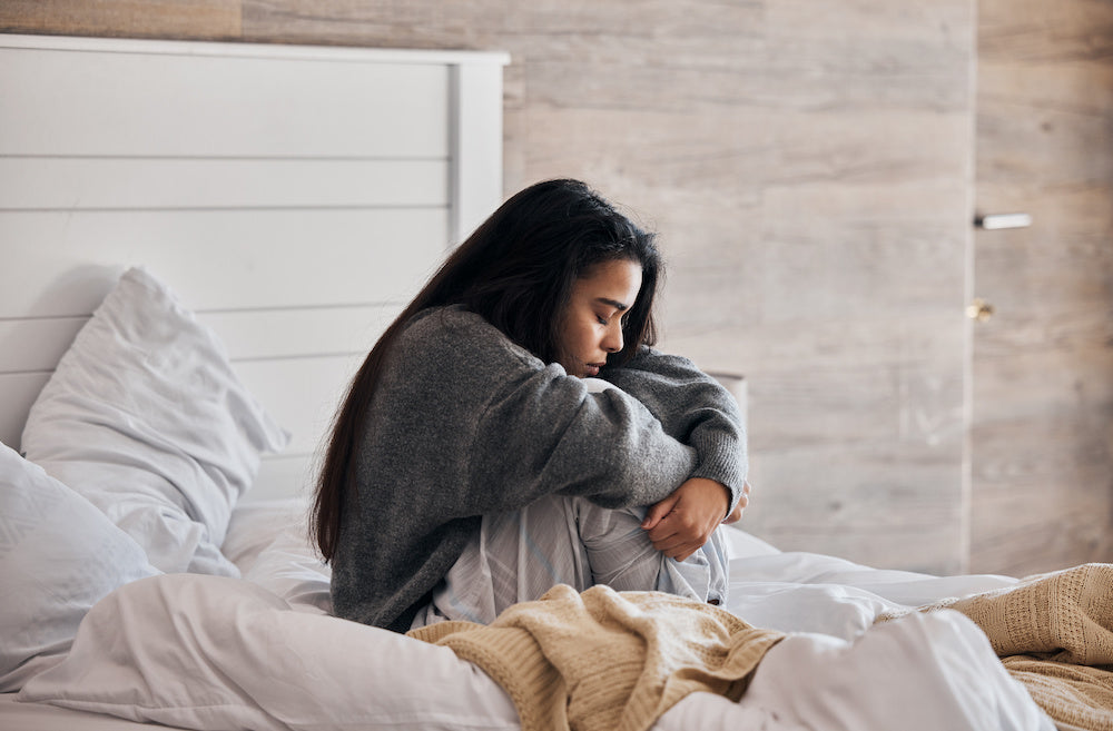 A color photo of a woman sitting on her bed experiencing disorienting morning anxiety