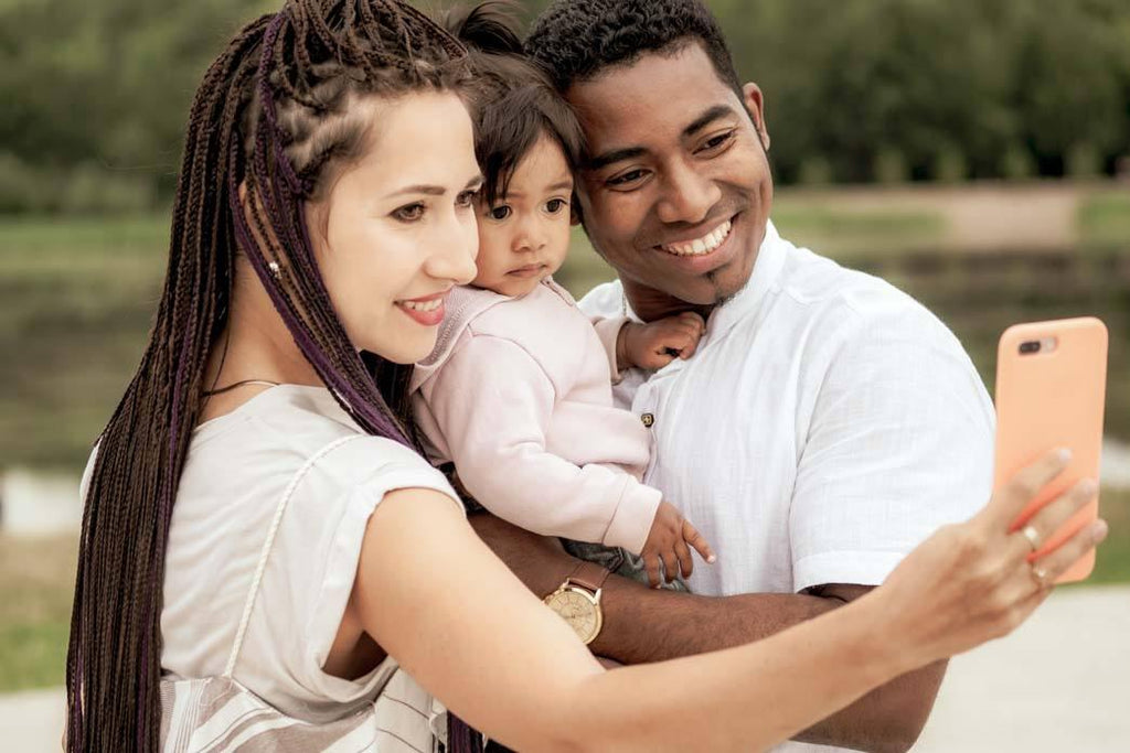 This is a photo of a happy young couple spending time with their daughter taking a family selfie. One parent is white with long black hair. The other parent is black with short hair.