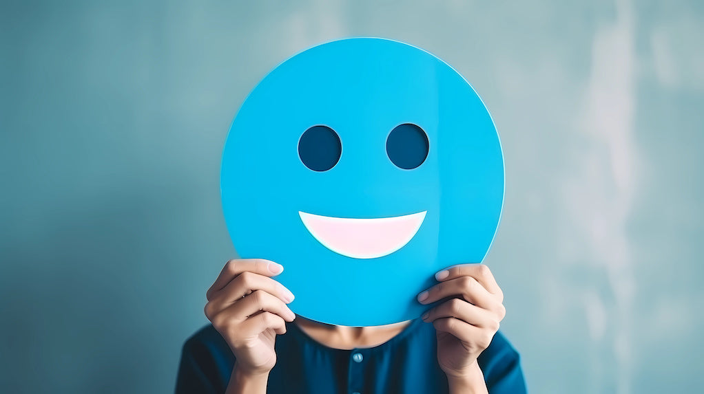 Improving your employees' mental health: a photo of a person holding blue paper cut happy smile face in front of their face.