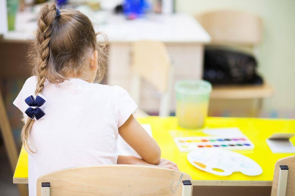 Childhood scoliosis surgery stories: a photo of girl at primary school. She is sitting at a yellow desk doing arts and crafts, her back facing the camera, her spine bent to the left.