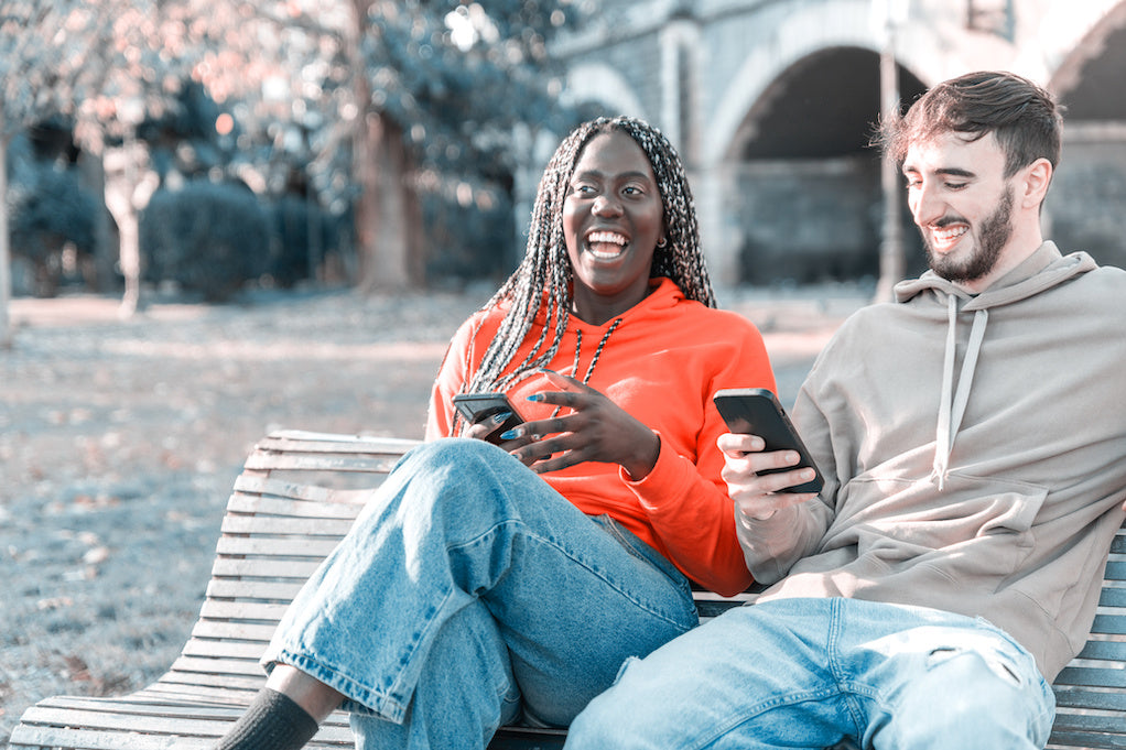 Two people laughing together while looking at a phone on a park bench.