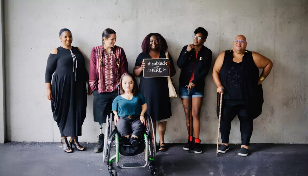 A diverse group of disabled people standing together, smiling confidently in front of a concrete wall. Some use mobility aids, and one holds a sign reading “disabled and here.”