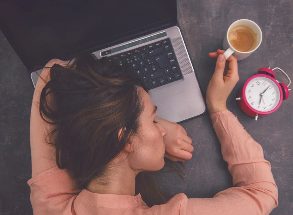 Person resting their head beside a laptop, holding a cup of coffee.