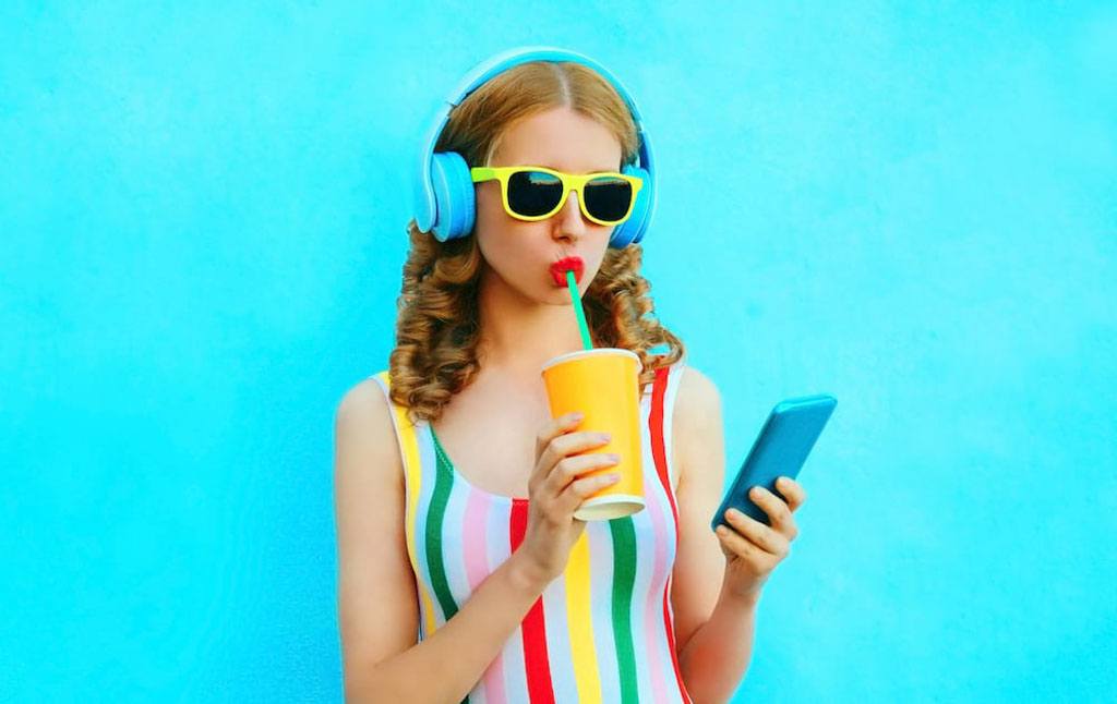 Image for article: How swiping right can help social anxiety ? Bright portrait of girl in bright clothing, drinking fruit juice, and holding a phone on colorful blue background