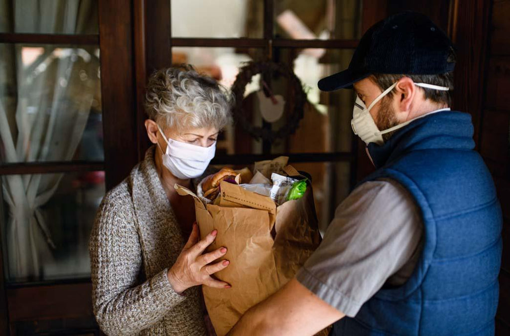 Costco personal shopper service: color photo of delivery person handing a paper grocery bag to a senior woman. Both of them are wearing face masks.