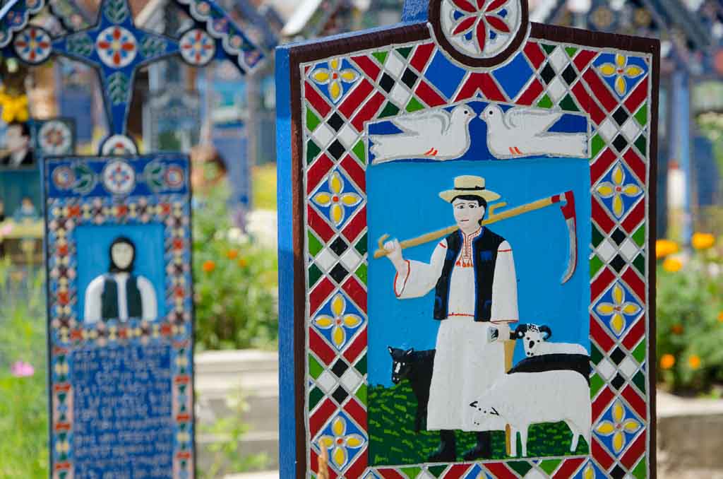 Romanian death traditions: a photo of the cheerful cemetery of Sapinta in Romania. A close up photo of a grave, complete with colorful painted wooden grave markers with a painting of a farmer and a sheep, a key part of Romanian death rituals.