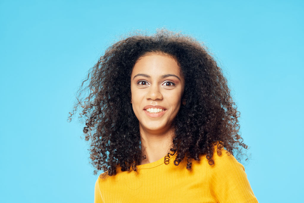 A photo of a woman with smiling depression. The woman is smiling at the camera, with a wide-open smile. She has long flowing black curly hair and is wearing a yellow jumper.