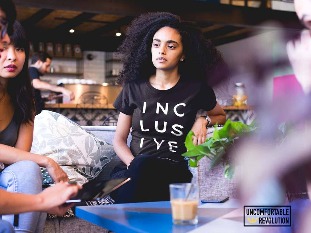 Benefits of emotional intelligence in the workplace in a cafe. Work colleagues are talking to each other. The person in the center has long wavy hair and is wearing a statement black t-shirt with the word inclusive is printed on it.