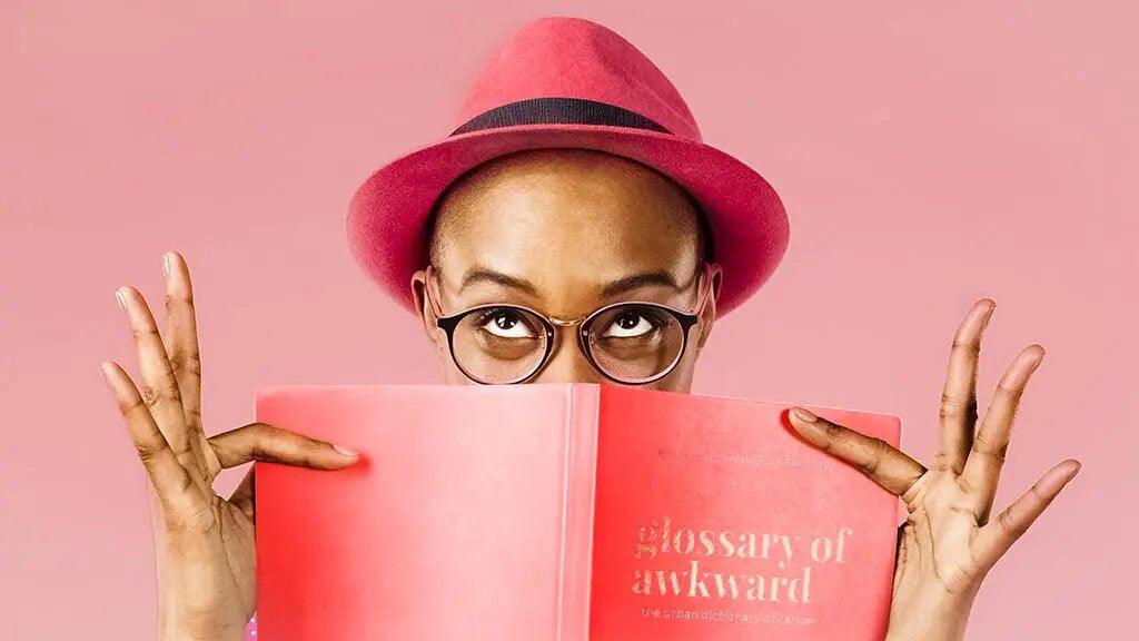Black woman with shaved head and pink hat. She is holding one of the funny books for cancer patients over her mouth while looking up cheekily through glasses. The title of the book is Glossary of Awkward.