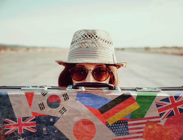 Image for article having a panic attack at airport. Hipster girl in a hat and sunglasses hides half her face behind a vintage suitcase with stamps flags of different country