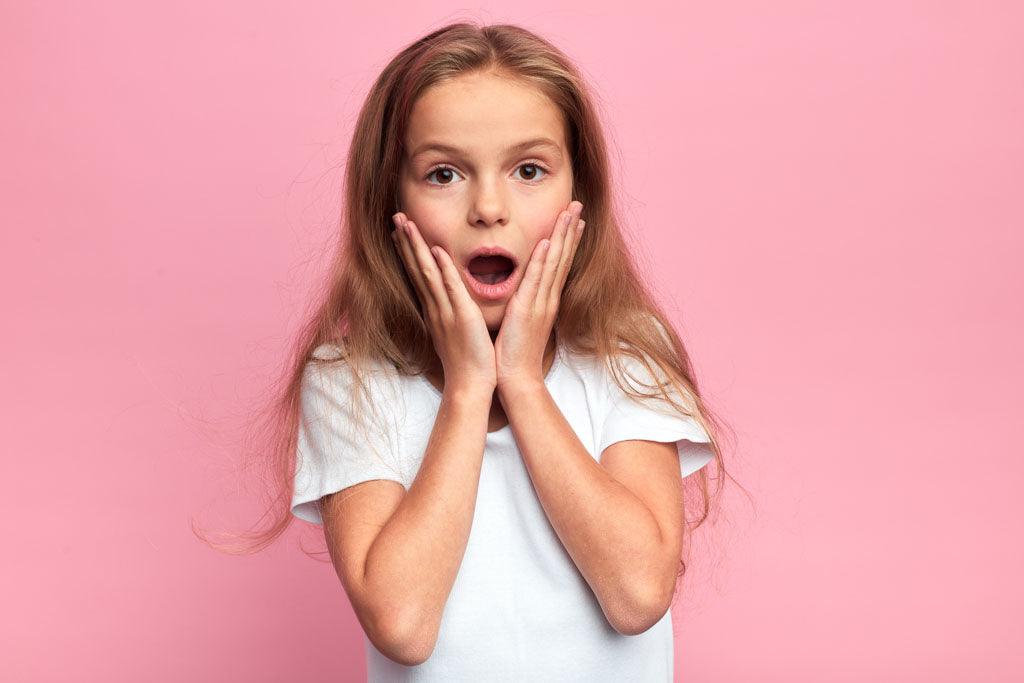 Questions to ask someone in a wheelchair: A close up portrait of a surprised child in white t-shirt with their mouth open and both hands on their cheeks. They are standing against a pink wall.